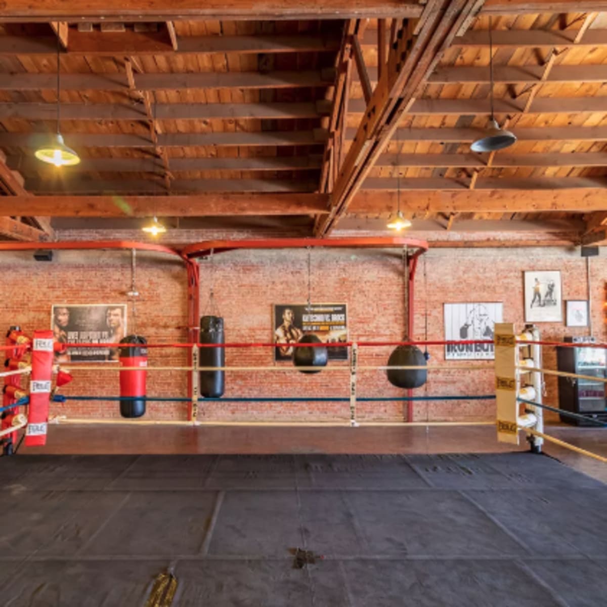 A boxing gym featuring a ring surrounded by heavy bags and adorned with vintage boxing posters on the brick walls.