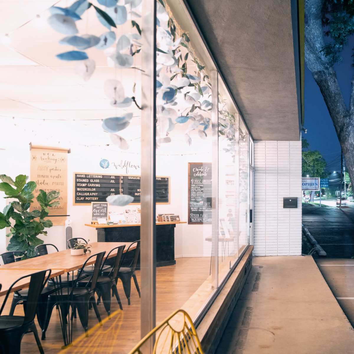 A bright café interior with a large communal table, plants, and a menu board visible through expansive windows at night.