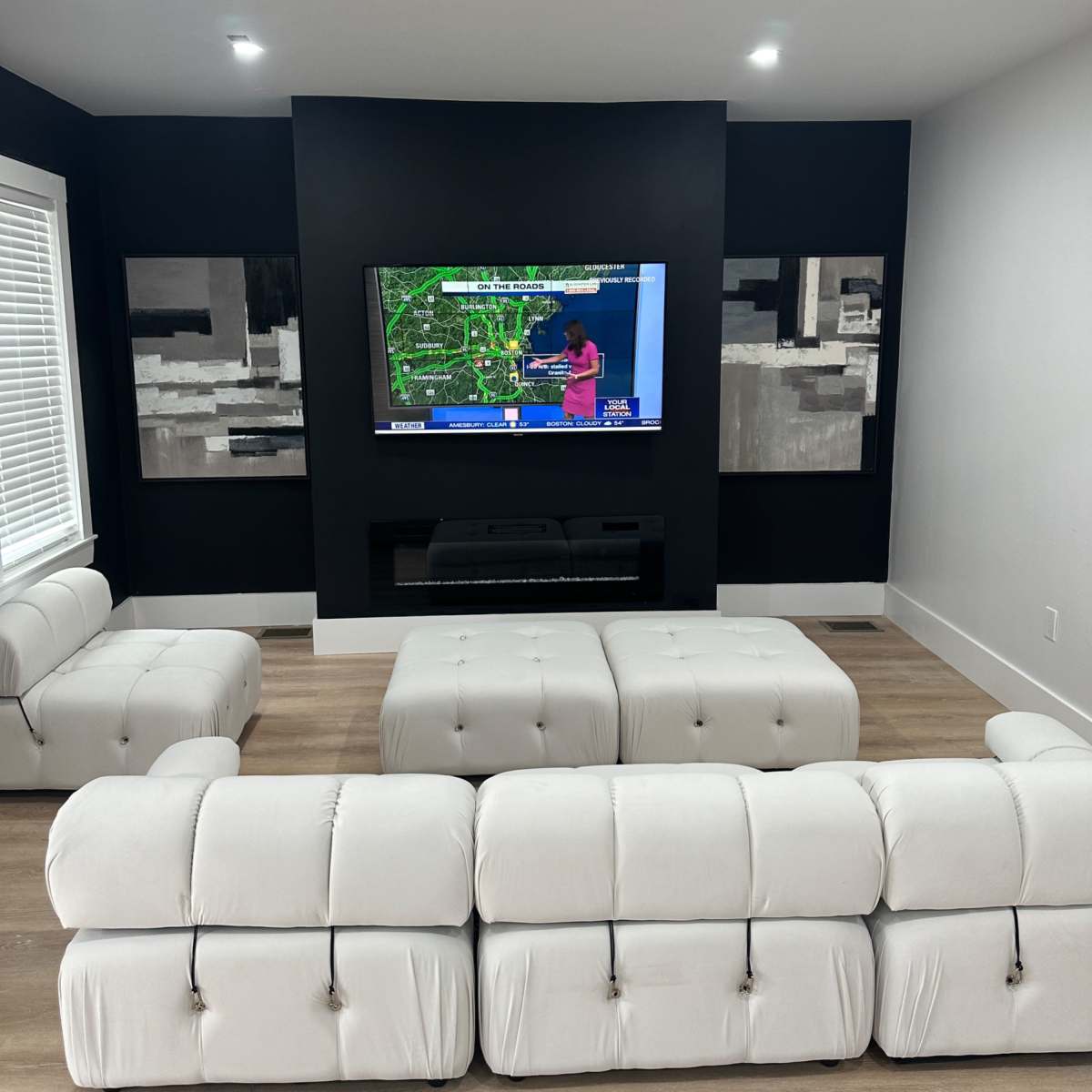 A modern living room with a white sectional sofa facing a mounted TV on a black wall, flanked by two abstract art pieces.