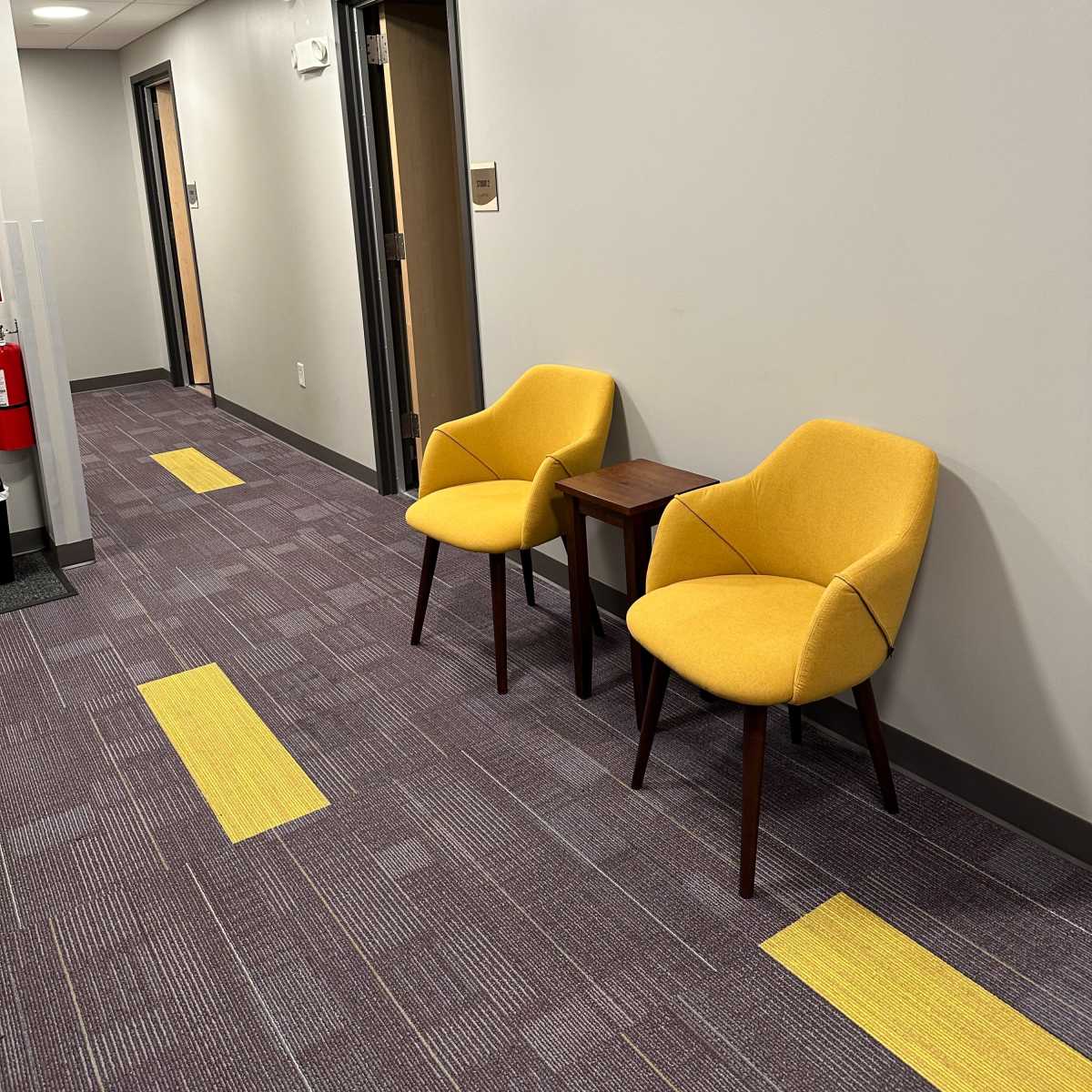 A hallway with two yellow chairs and a small table positioned against a wall, with doors along the corridor.