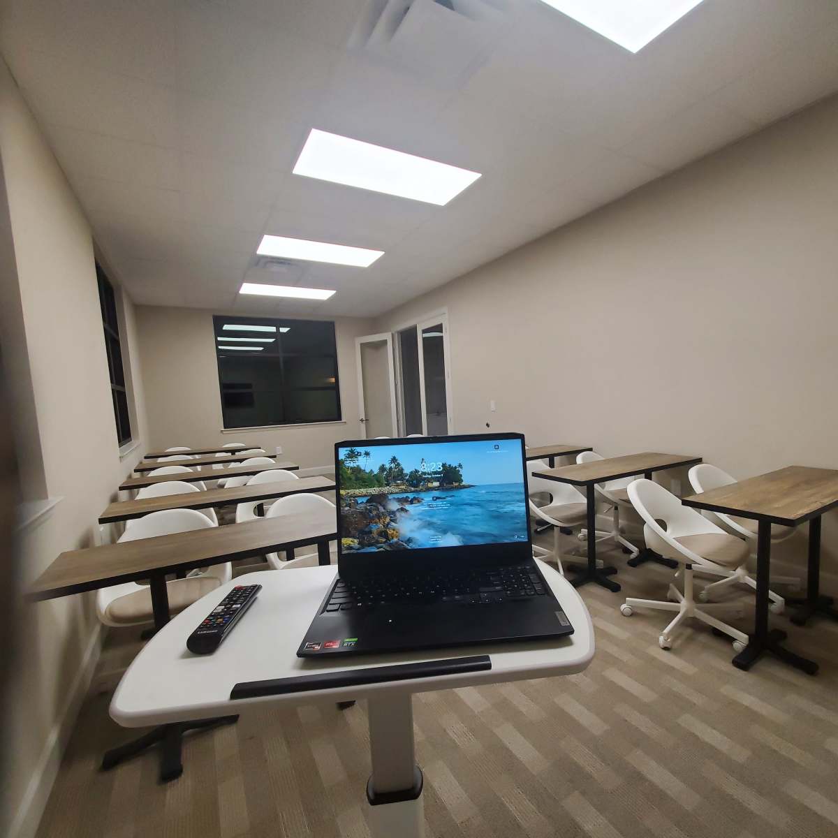 A laptop displaying a beach scene is set on a desk in a nearly empty classroom with rows of chairs and tables.