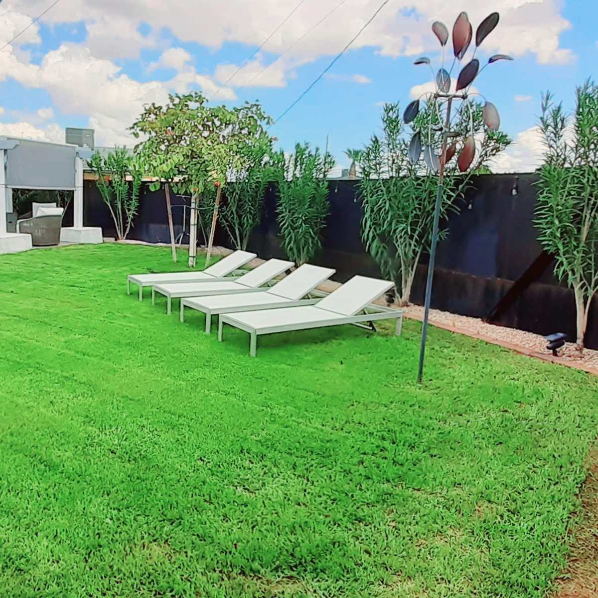 A well-maintained lawn with three white lounge chairs, a covered patio area, and decorative plants along the perimeter.