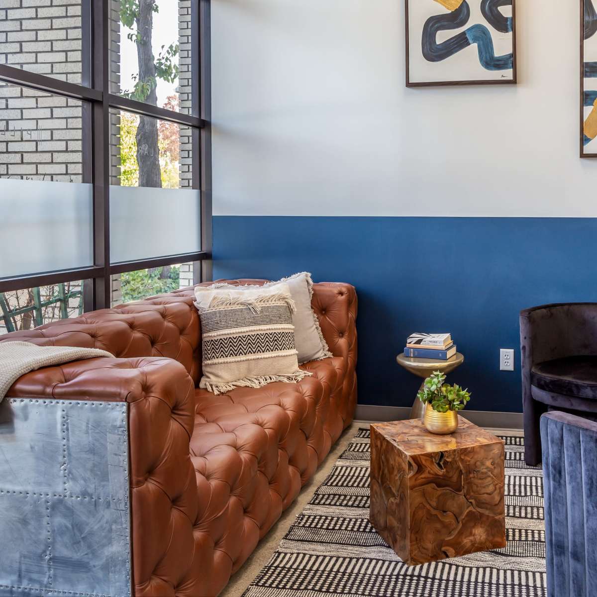 A modern seating area featuring a brown leather sofa, two black velvet chairs, a wooden side table, and abstract artwork on the walls.