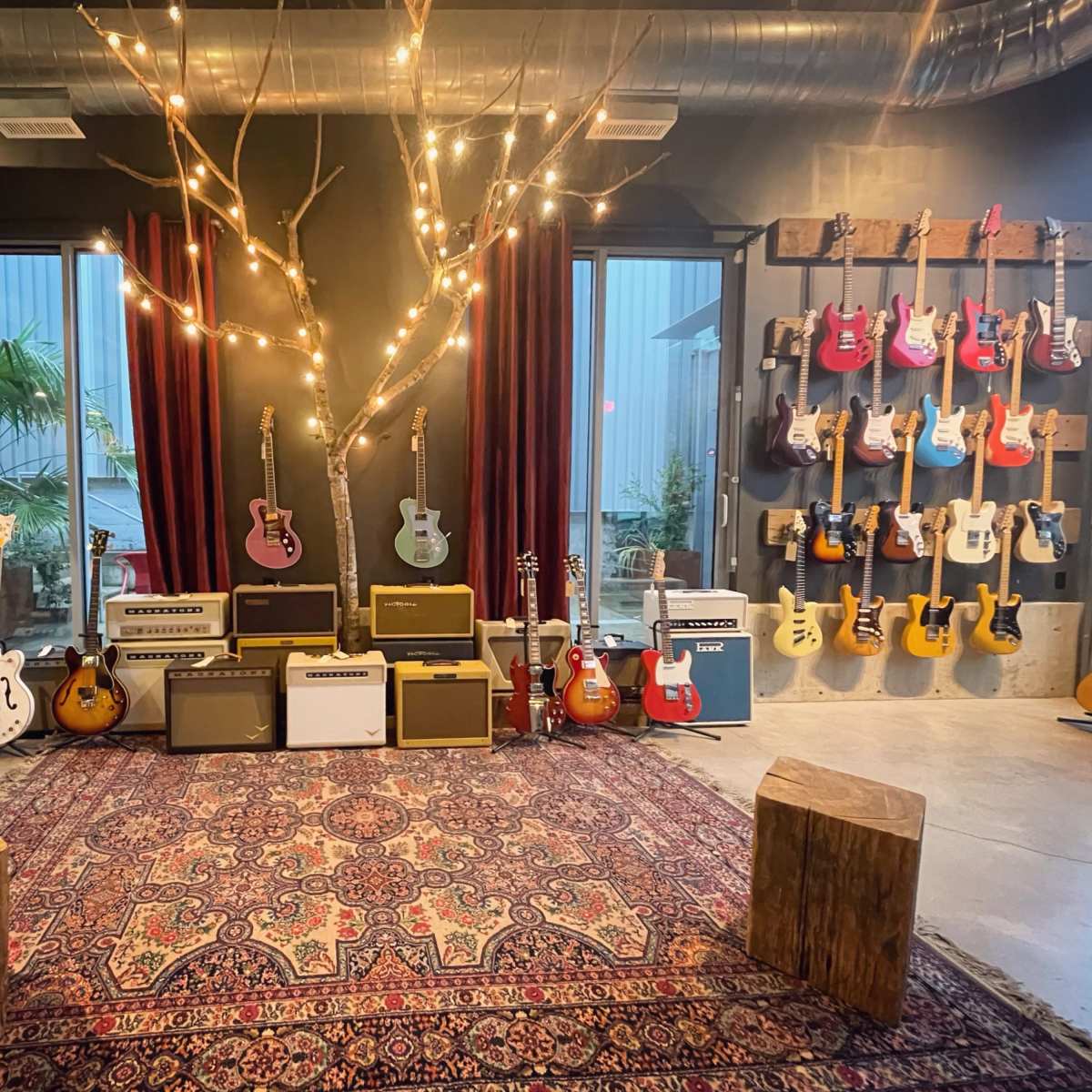 A music studio with various guitars displayed on the walls, amplifier stacks in the center, and wooden stools on a patterned rug.
