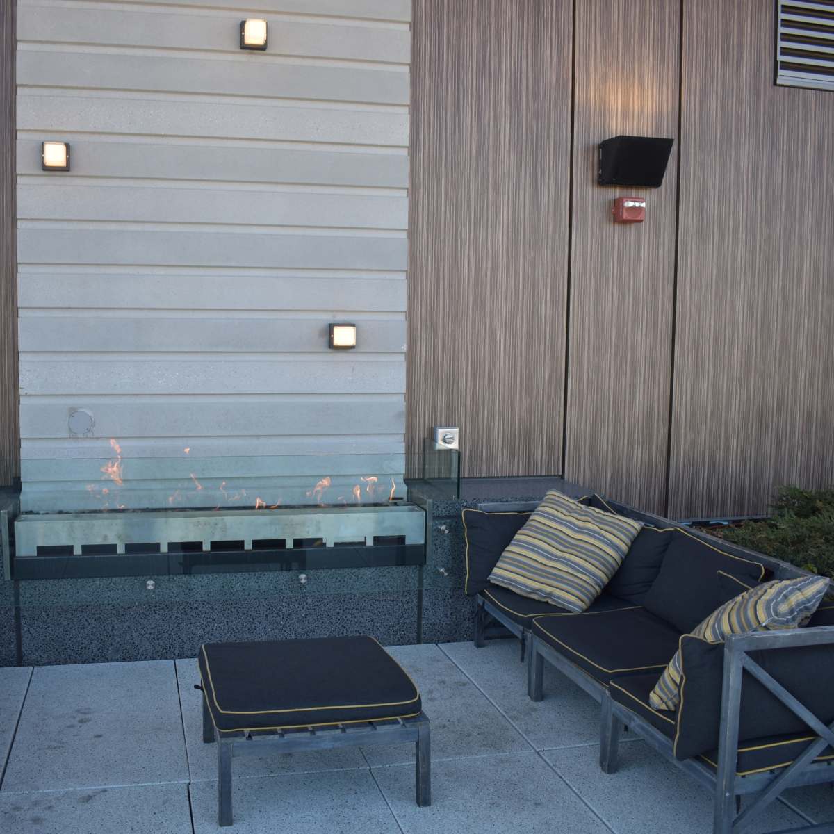 A seating area featuring gray outdoor furniture arranged around a linear fire feature against a wooden wall.