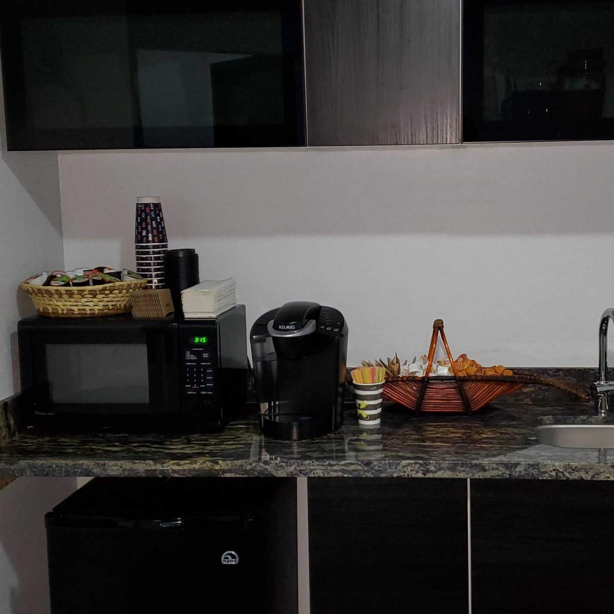 A kitchen corner with a microwave, coffee maker, sink, and a basket of snacks on a countertop.