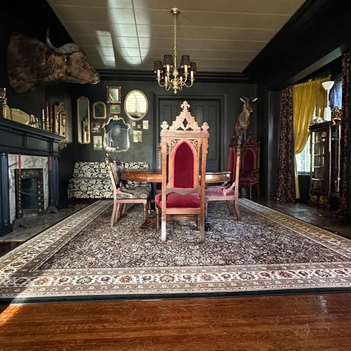 A dimly lit dining room featuring an intricately designed wooden table surrounded by high-backed chairs, a large area rug, and various decorative elements on the walls.