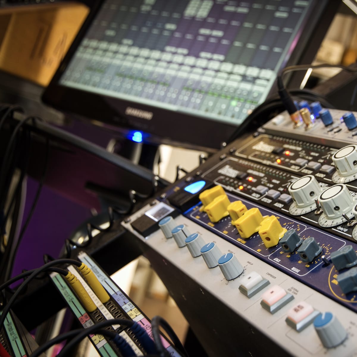 A close-up of audio mixing equipment featuring knobs and sliders alongside a computer screen displaying digital audio tracks.