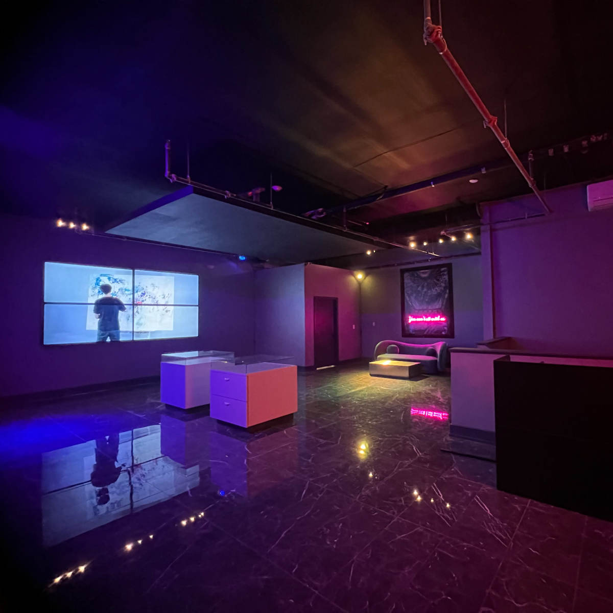 The interior of a dimly lit room features a large screen displaying a figure, modern furniture, and colorful lighting effects.