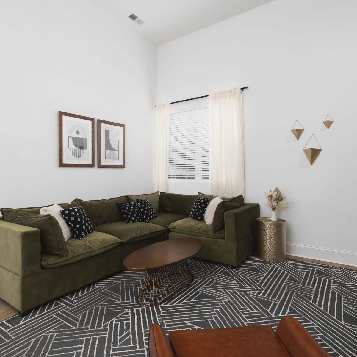 A green sectional sofa is arranged in a bright living room featuring a round coffee table, two framed artworks on the wall, and decorative items on a shelf.