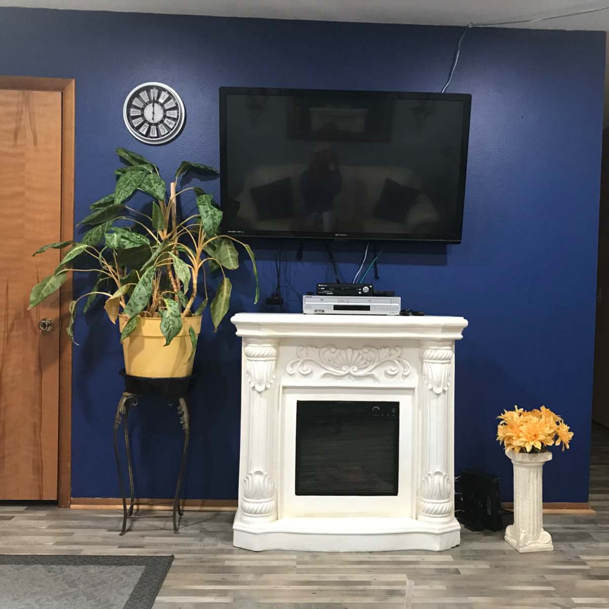 A living room with a blue wall, a mounted television above a white fireplace, a potted plant in a yellow planter, and a decorative pedestal with yellow flowers.