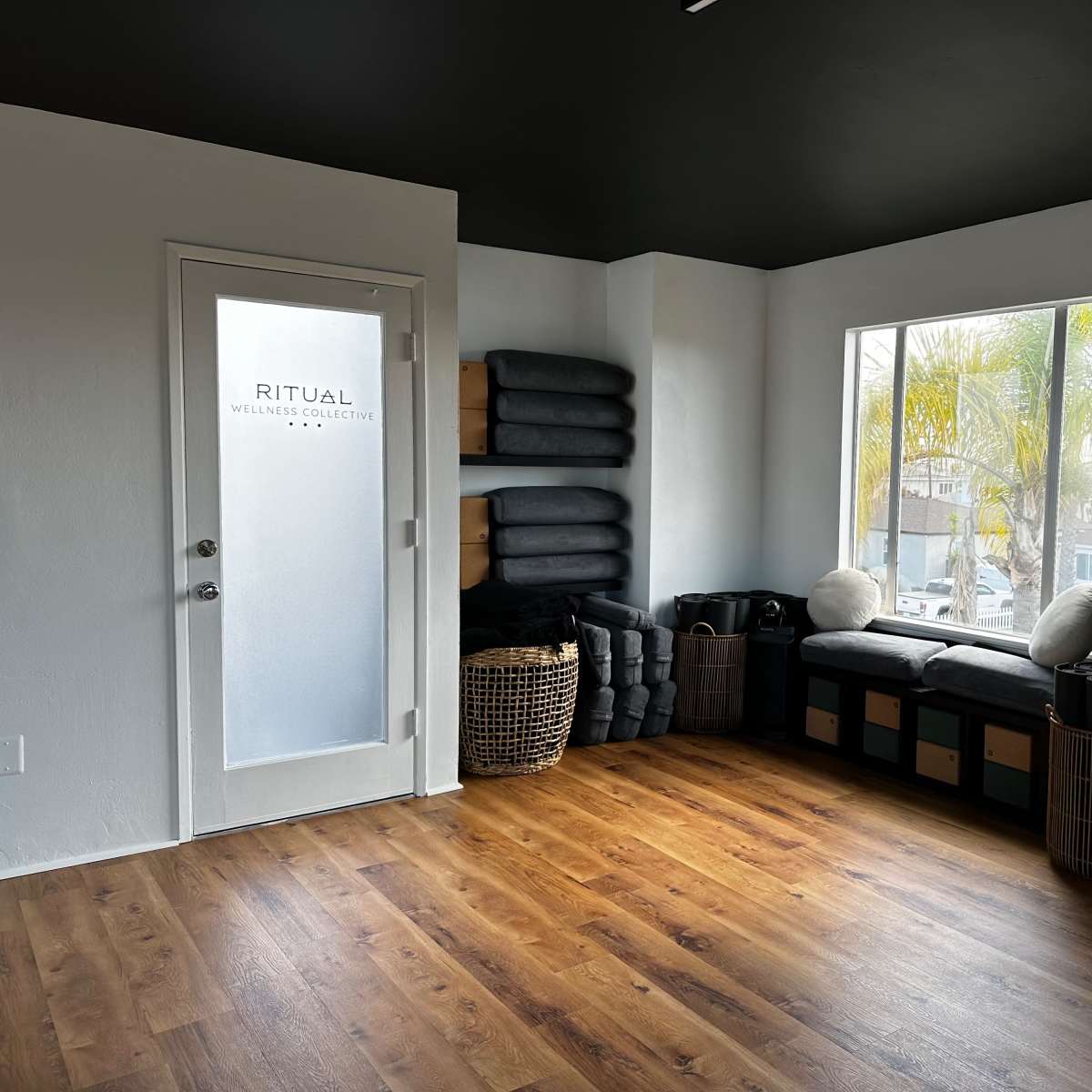 A minimalist interior room with a window seat, stacked blankets, and a glass door labeled "RITUAL WELLNESS COLLECTIVE."