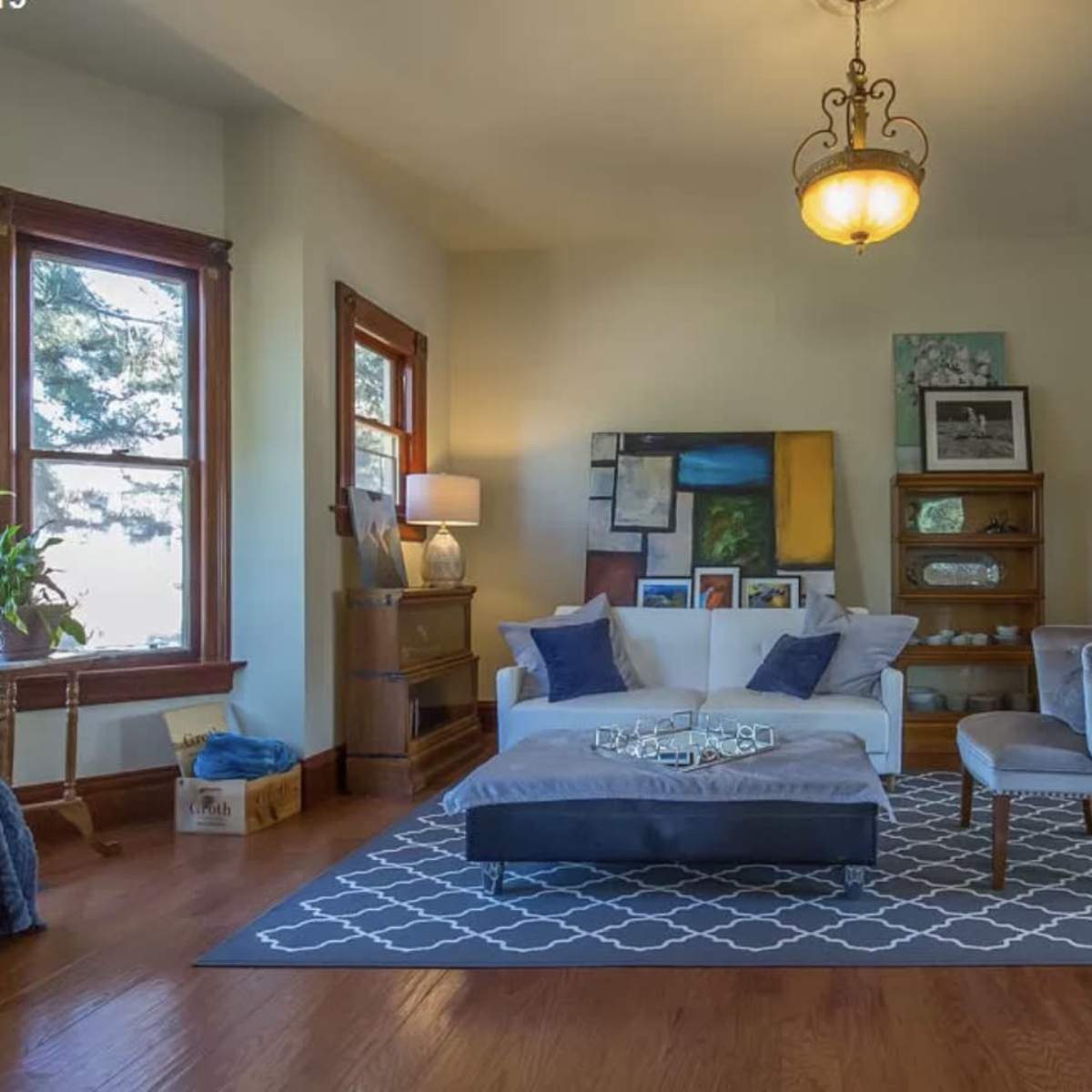 A brightly lit living room features a white sofa with blue cushions, a grey ottoman, and wooden furniture, accented by large windows and artwork on the walls.