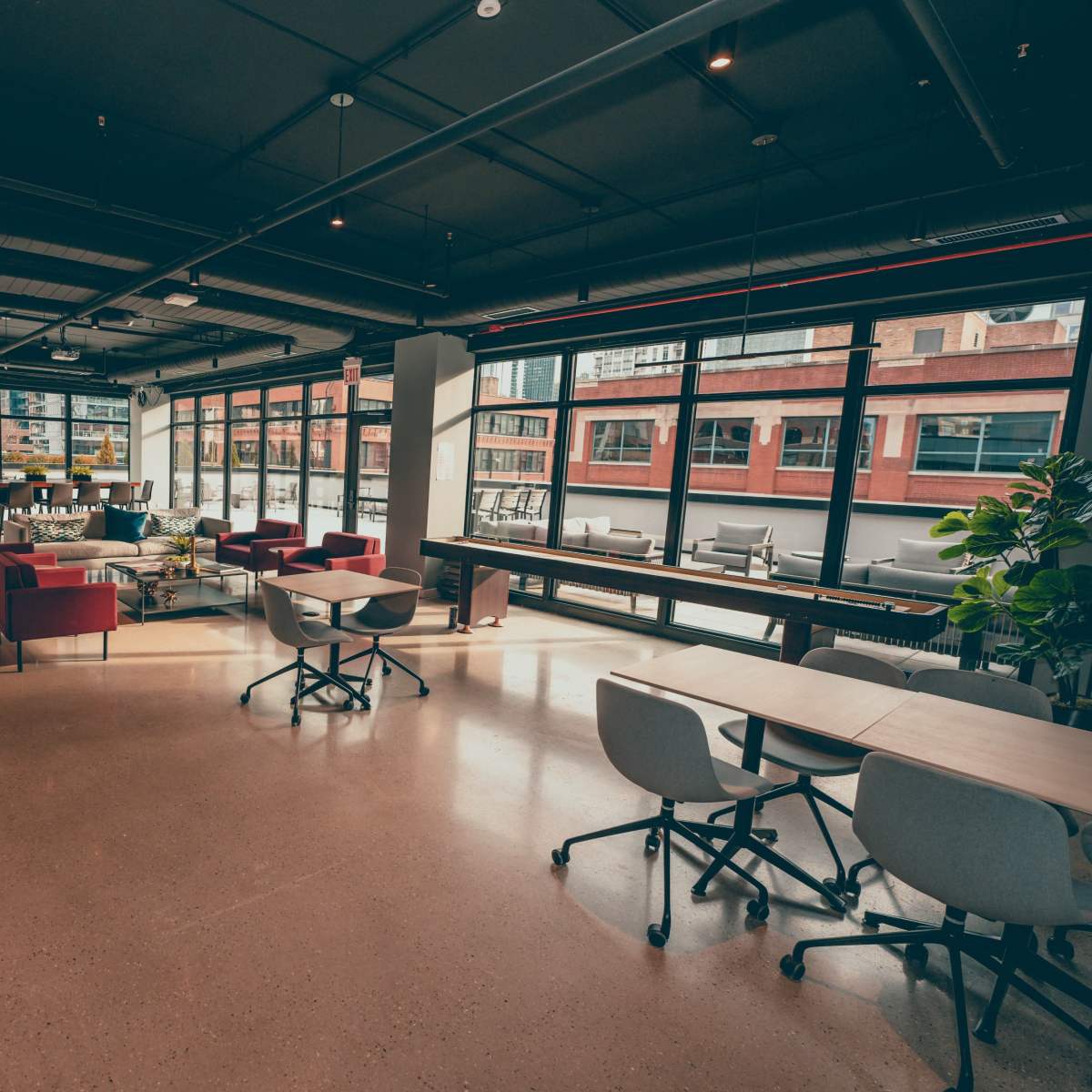 A modern office space with large windows, multiple seating areas, and various tables arranged throughout.