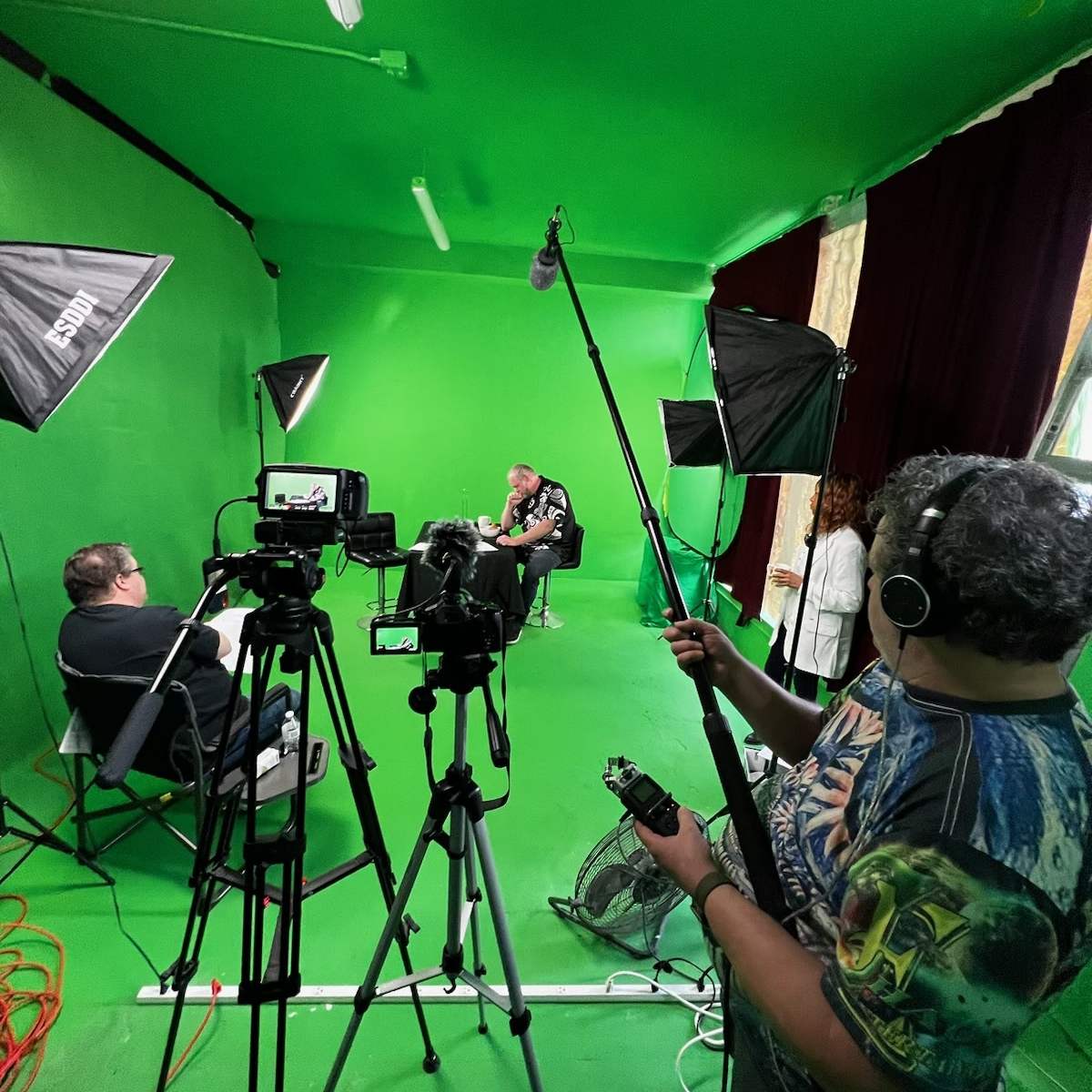 A film crew sets up equipment in a green-screen studio, with cameras, lights, and a microphone being positioned for a shoot.