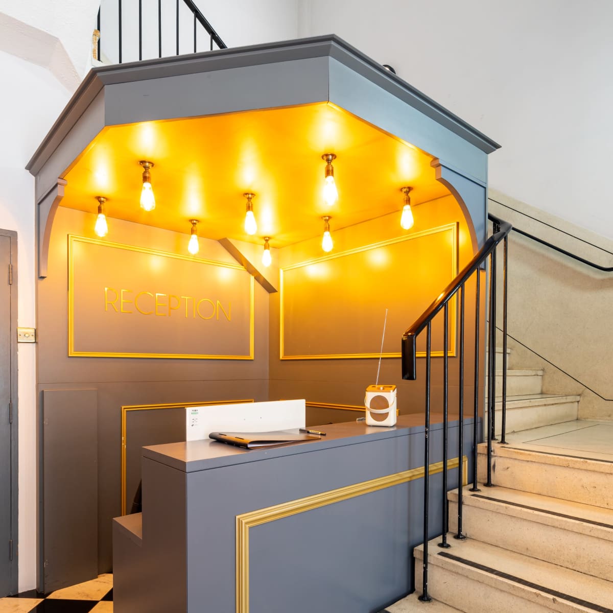 A modern reception desk with yellow lighting and a staircase leading up beside it, situated in a space with black and white flooring.