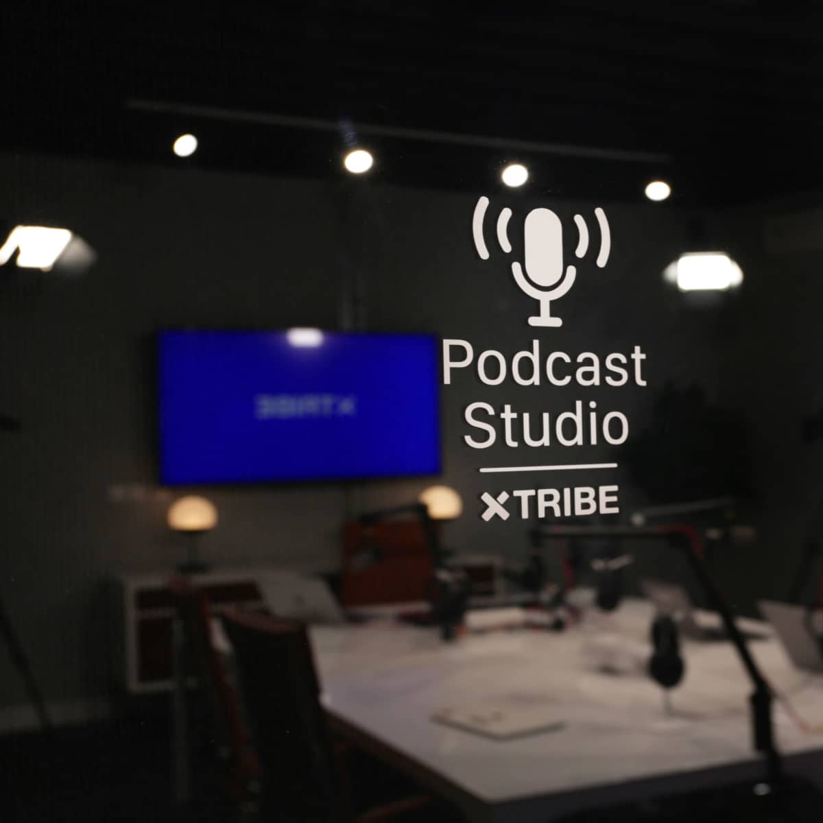 A glass door with the words "Podcast Studio" and "TRIBE" etched on it, showing a modern recording space with microphones and a large screen visible inside.