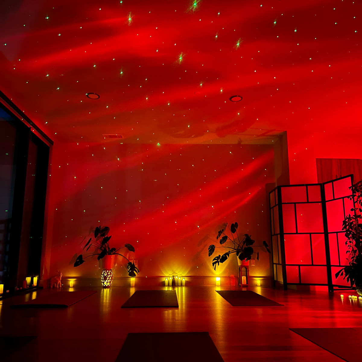 A dimly lit yoga studio with red lighting, star-like projections on the walls, and plants placed around the space.