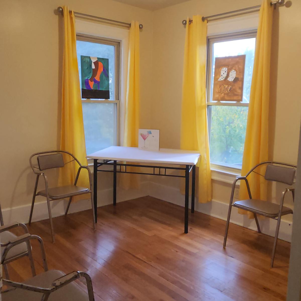 A small room with two windows, yellow curtains, a white table in the center, and three metal chairs arranged around it.