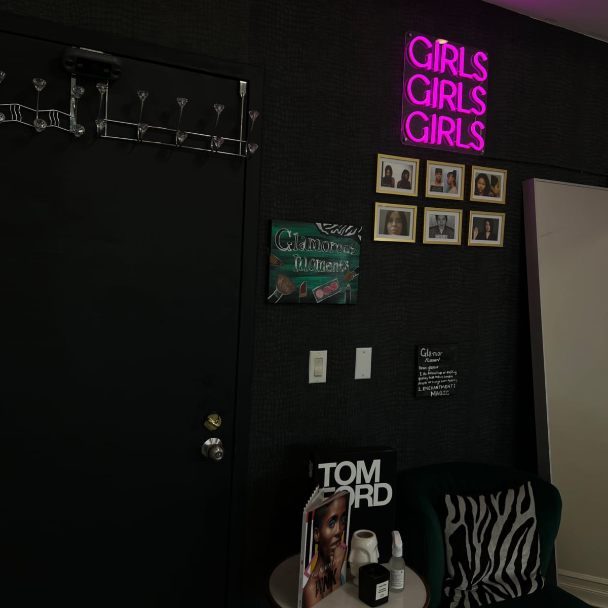 A dark wall with a neon sign that reads "GIRLS GIRLS GIRLS," a mirror, framed photographs, and a green chair displaying a magazine and skincare products.
