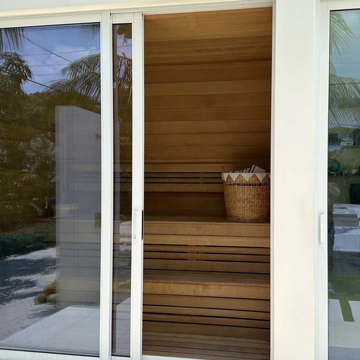 A modern sauna made of wood, located beside a large outdoor pool area surrounded by palm trees.