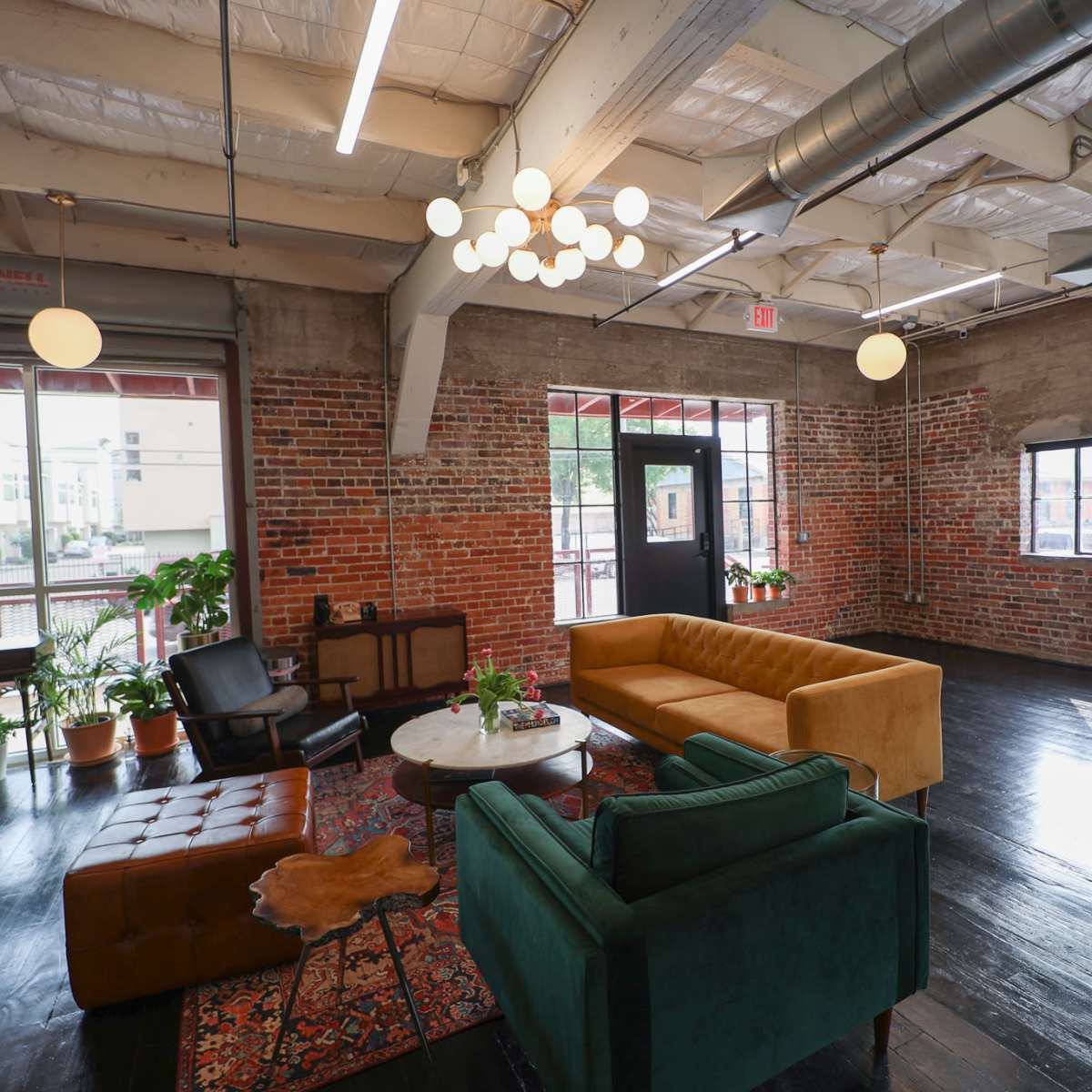 A spacious, loft-style room with exposed brick walls, modern light fixtures, and a mix of seating arrangements including a mustard-colored sofa, a green armchair, and a small coffee table surrounded by plants.
