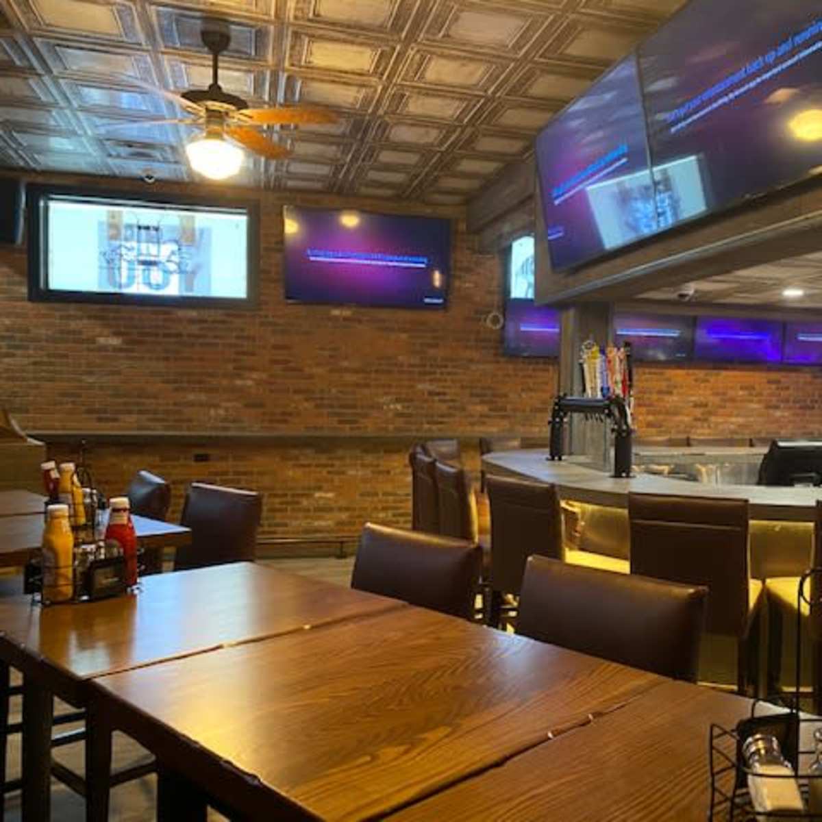 The interior of a bar or restaurant with wooden tables, leather chairs, a circular bar counter, and multiple screens displaying content on the walls.