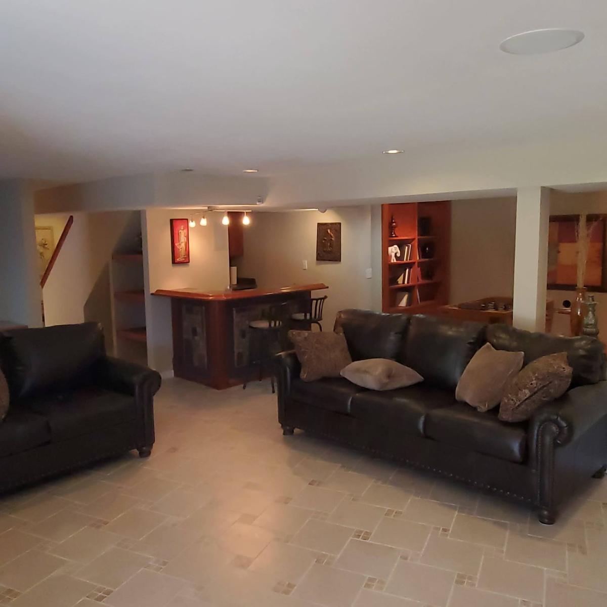 A finished basement with two leather sofas, a bar area, and a shelf filled with books.