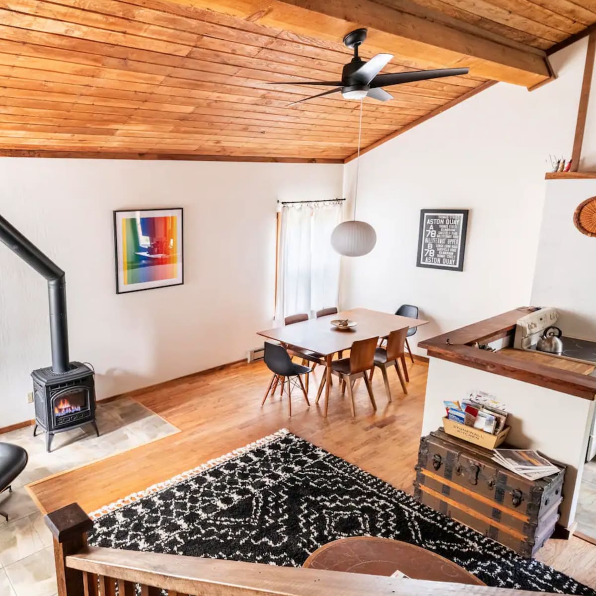 A spacious living area features a metal wood stove, a dining table, and large windows with light curtains.
