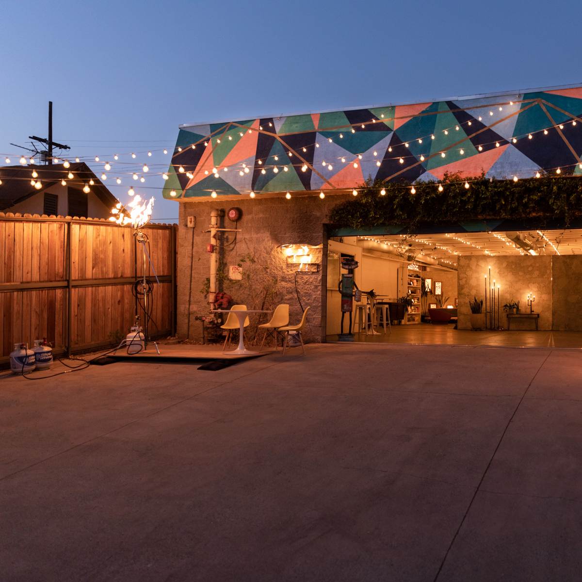 A garage with a colorful geometric mural, string lights overhead, and a spacious driveway leading to an open door.