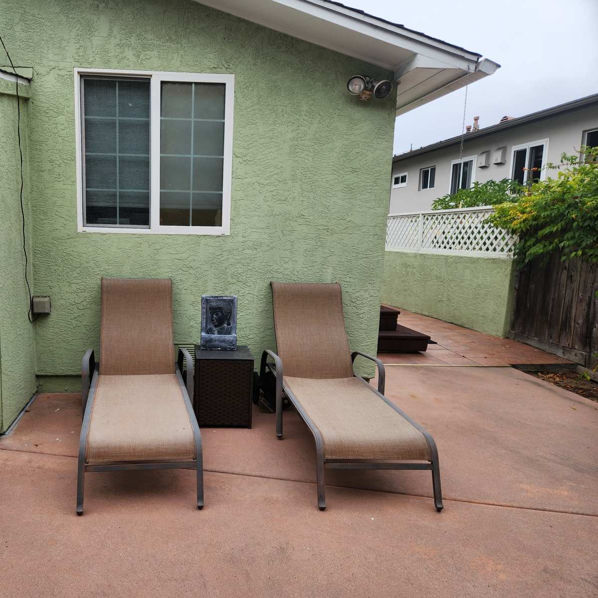 Two lounge chairs positioned next to a gray stone fountain against a green exterior wall of a house.