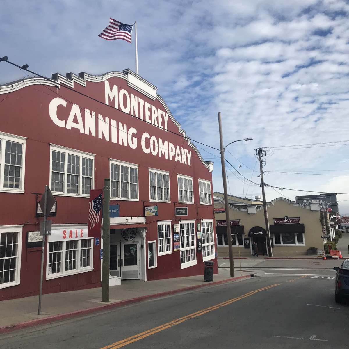 The red and white exterior of the Monterey Canning Company building, with an American flag flying atop and shops lining the street in the background.