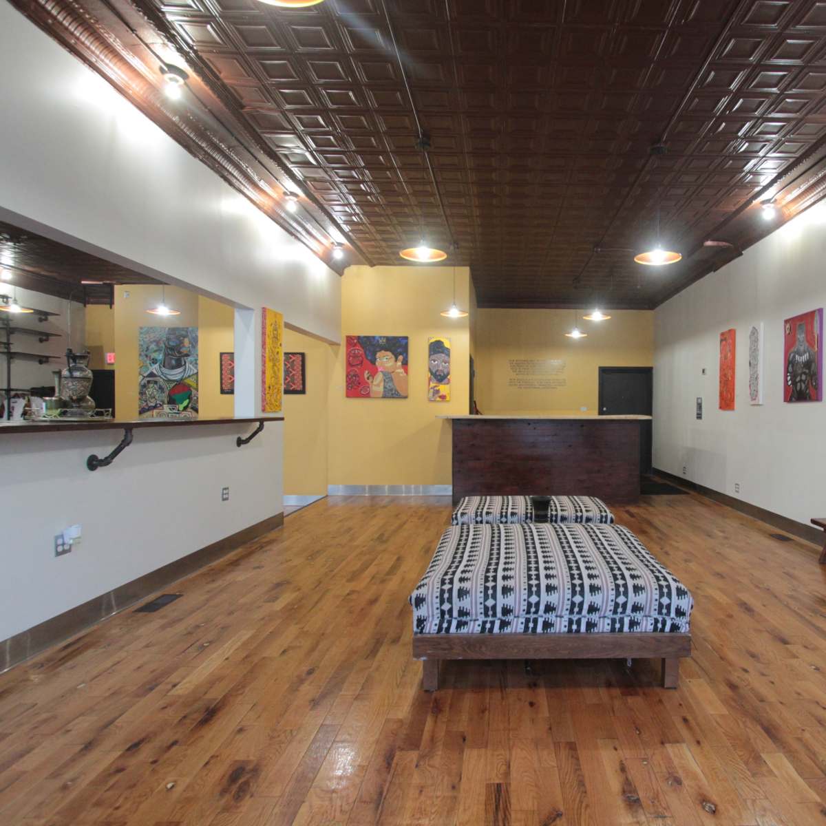 A spacious interior of a room with wooden floors, a patterned mattress on a wooden platform, a reception area on the left, and colorful artwork displayed on the walls.
