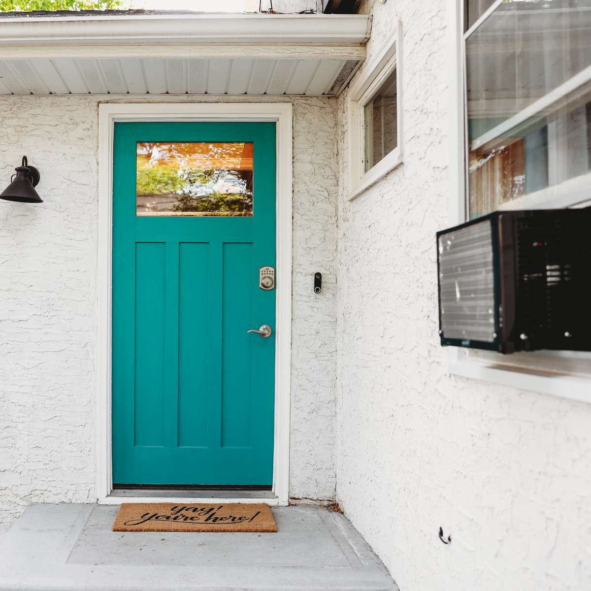A turquoise front door with a rectangular window is set against a white textured wall, accompanied by a doormat and an air conditioning unit beside it.