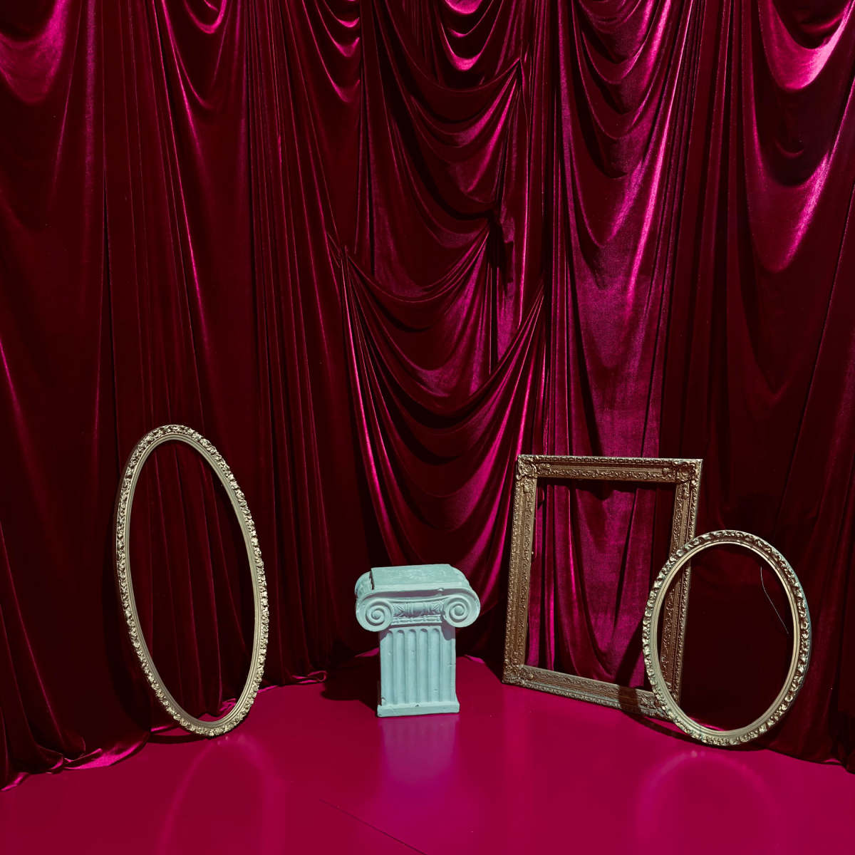The scene features a pink floor with a central turquoise column and two ornate frames, set against a backdrop of deep red velvet drapery.