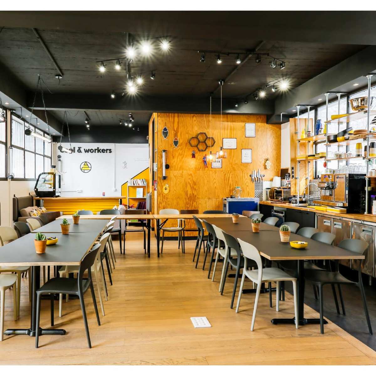 A modern café interior with wooden accents, long tables arranged in a communal setting, and a well-stocked beverage counter.
