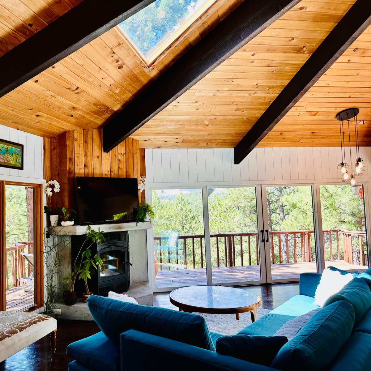 A bright, spacious living room with a wooden ceiling, large windows overlooking a forested area, and a modern fireplace.