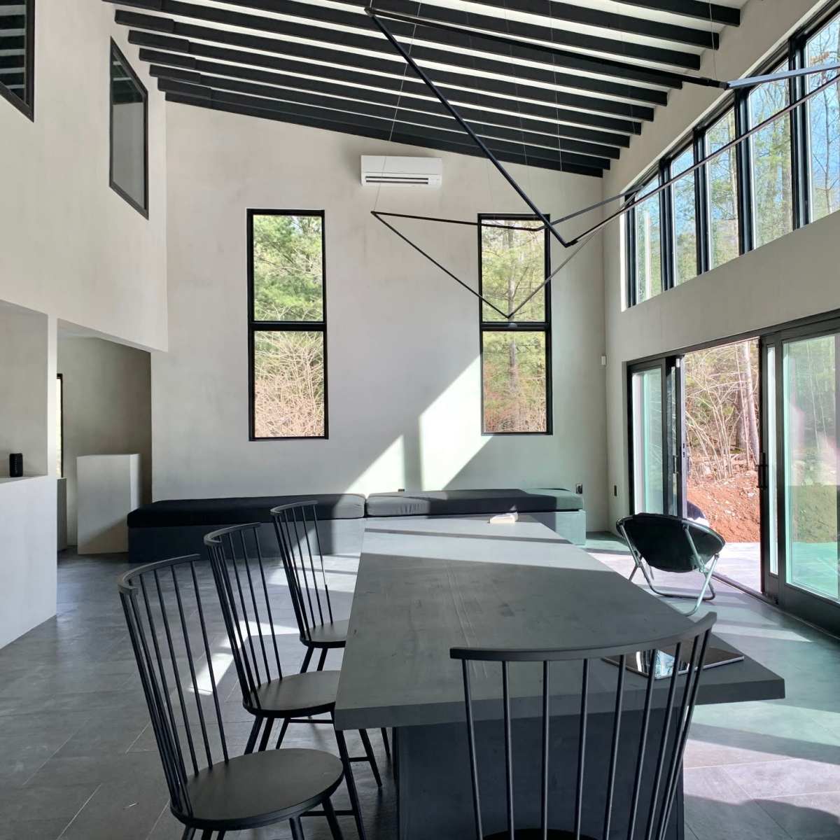 The interior of a modern, open-plan dining area features a large table surrounded by black chairs, with tall windows that allow sunlight to enter and a view of the outdoors.