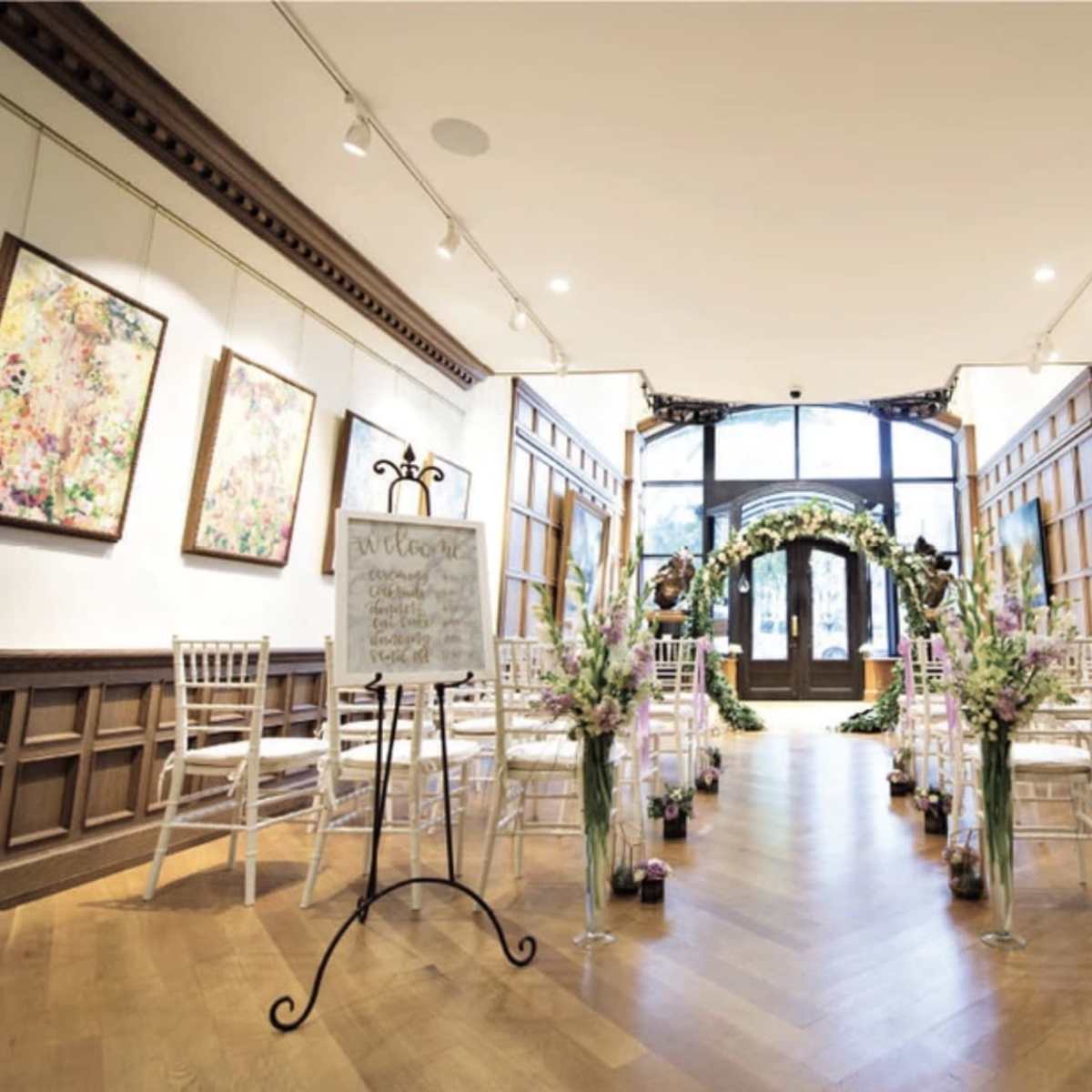 A wedding ceremony setup features rows of white chairs facing an ornate arch adorned with greenery and flowers in a tastefully decorated gallery.