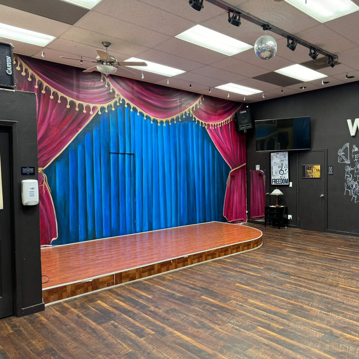 A stage with a blue curtain backdrop and red velvet drapes, set in a room featuring a wooden floor and a wall adorned with musical illustrations.