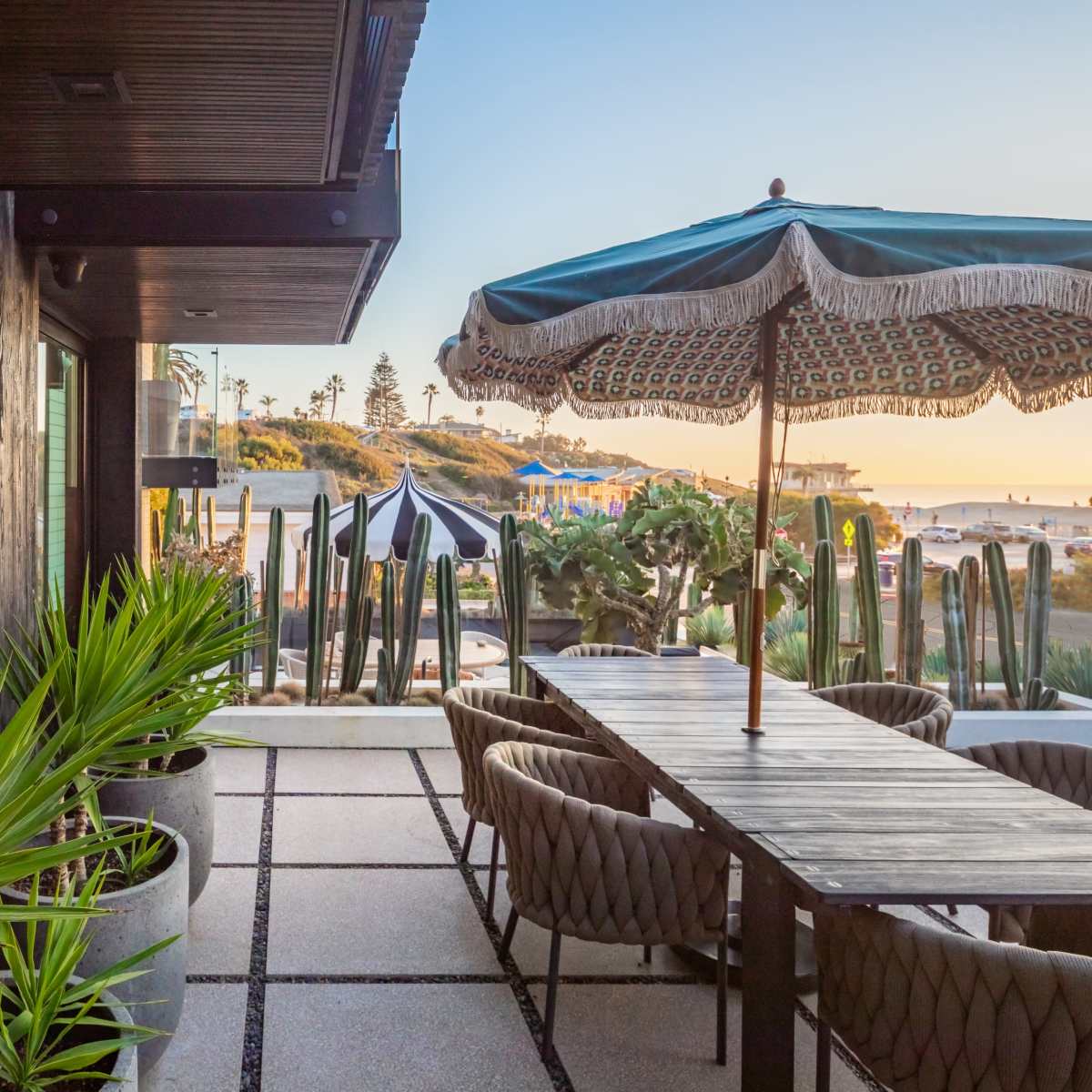 A patio area features a large table under a decorative umbrella, surrounded by potted plants, with a view of the beach in the background.