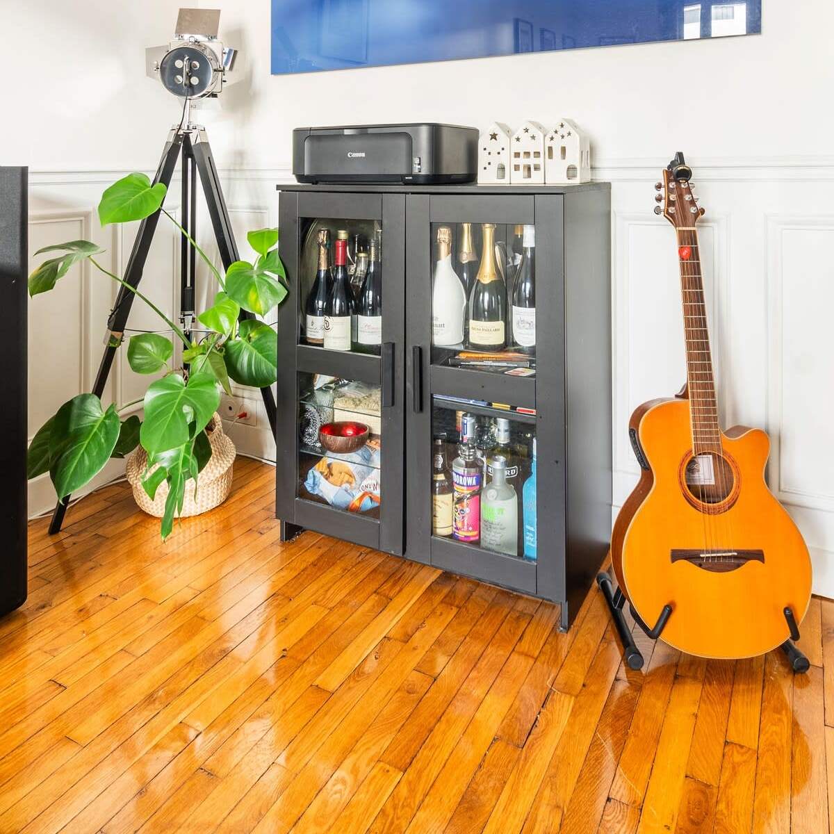 A black cabinet with glass doors displays various bottles, accompanied by a guitar, a large photograph of a lake scene, and a green plant in a corner of a room with wooden flooring.
