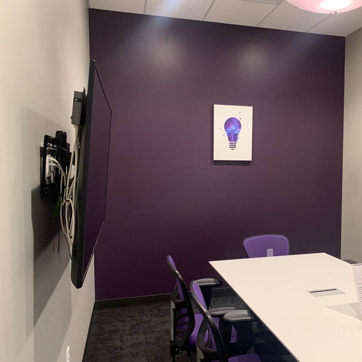 A small conference room features a white table, four purple chairs, a wall-mounted TV, and a light bulb-themed artwork on a deep purple wall.