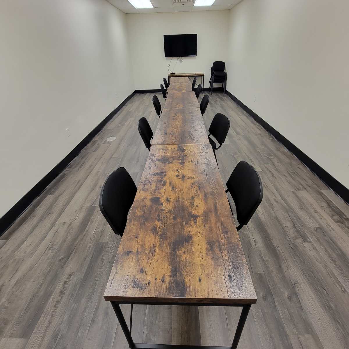 A long wooden table with black chairs is arranged in a meeting room, featuring a television mounted on the wall.