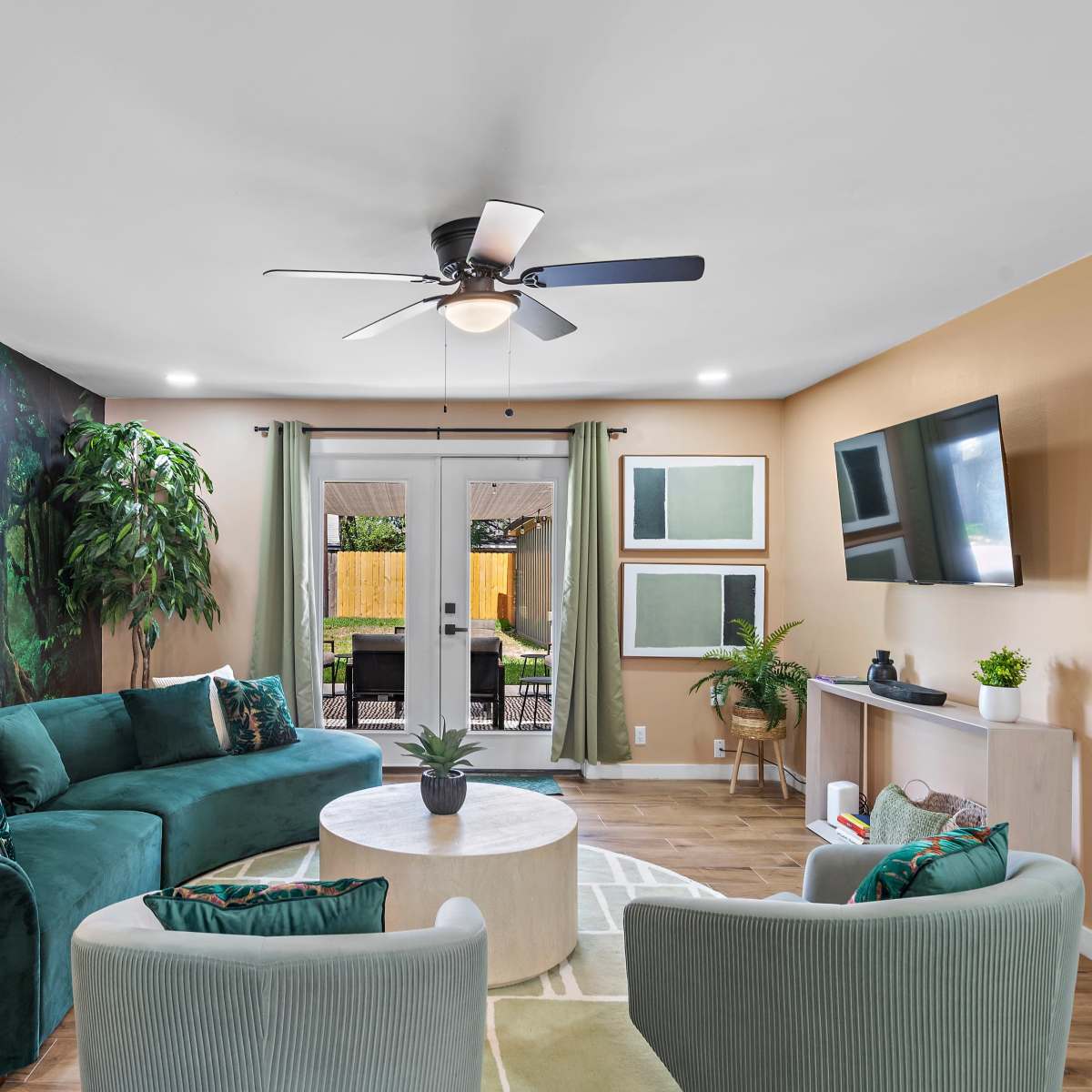 The living room features a green sectional sofa, a round coffee table, and large windows leading to a patio, adorned with a mural of trees on one wall.