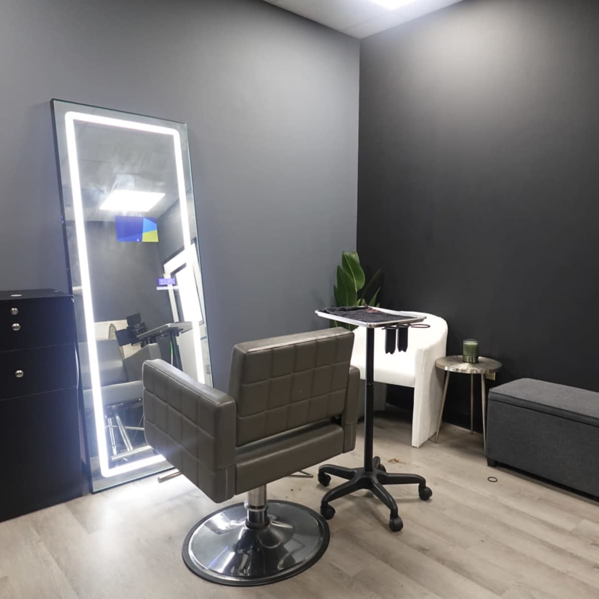 A modern hair salon interior featuring a stylist chair, a tall mirror, a small table with tools, and storage units against dark walls.