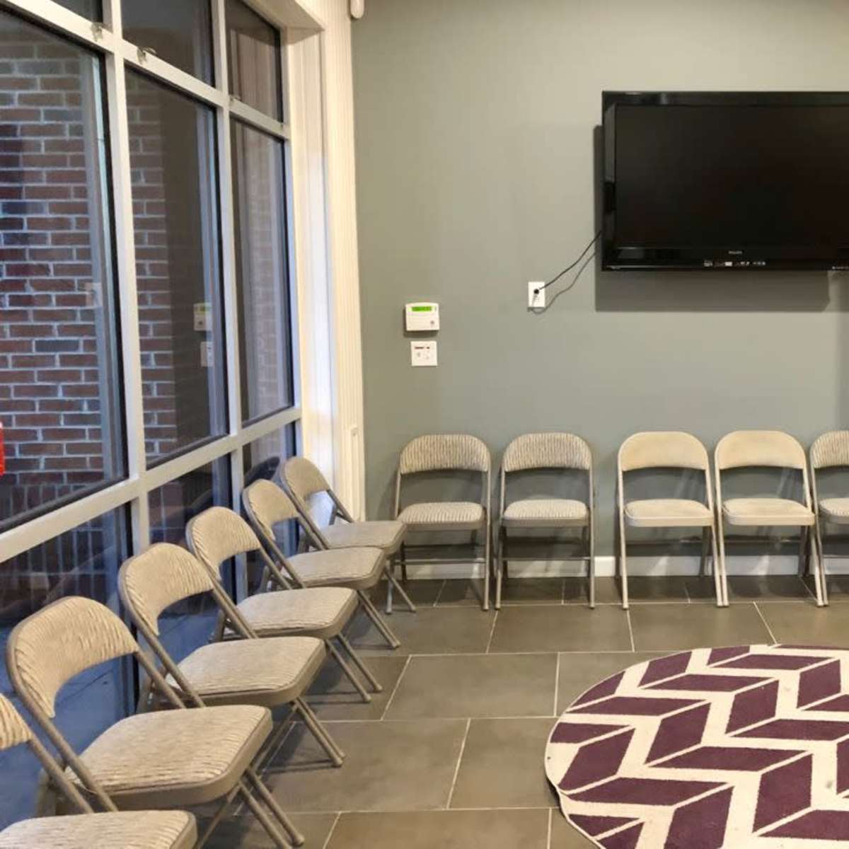 A room with a row of metal folding chairs facing a wall-mounted television, alongside a wooden media stand and a round purple rug on the tiled floor.