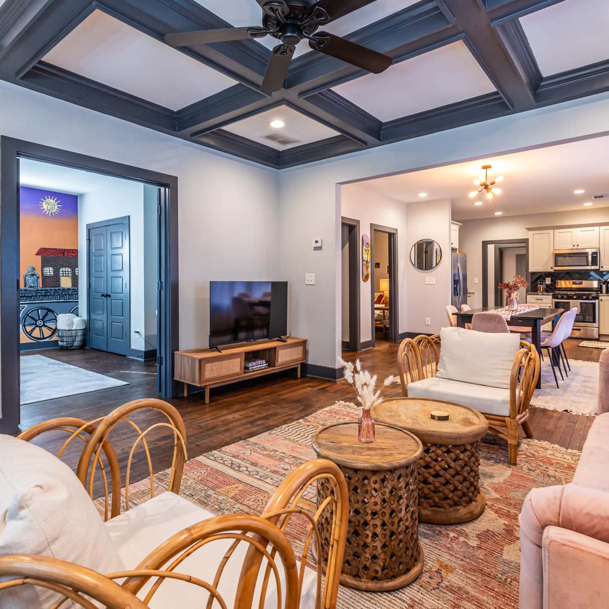 A living room with a coffered ceiling, wooden floor, and a mix of seating arrangements, including chairs and a sofa, leading to an adjacent dining area.
