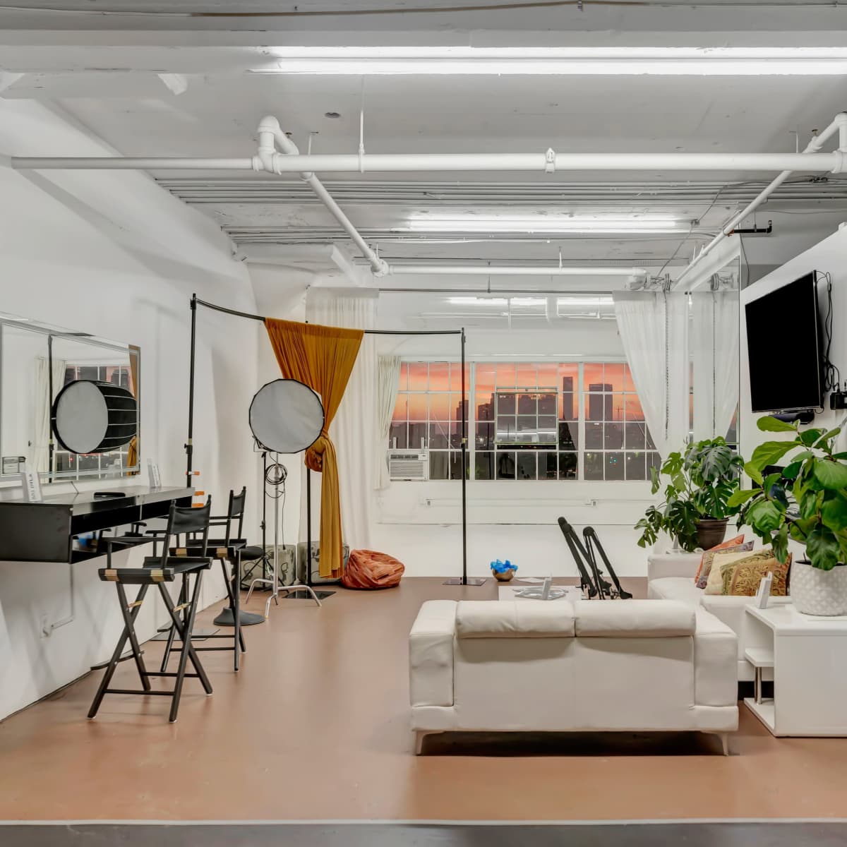 A bright, minimalist studio space with a sofa, makeup station, and large windows revealing a sunset.