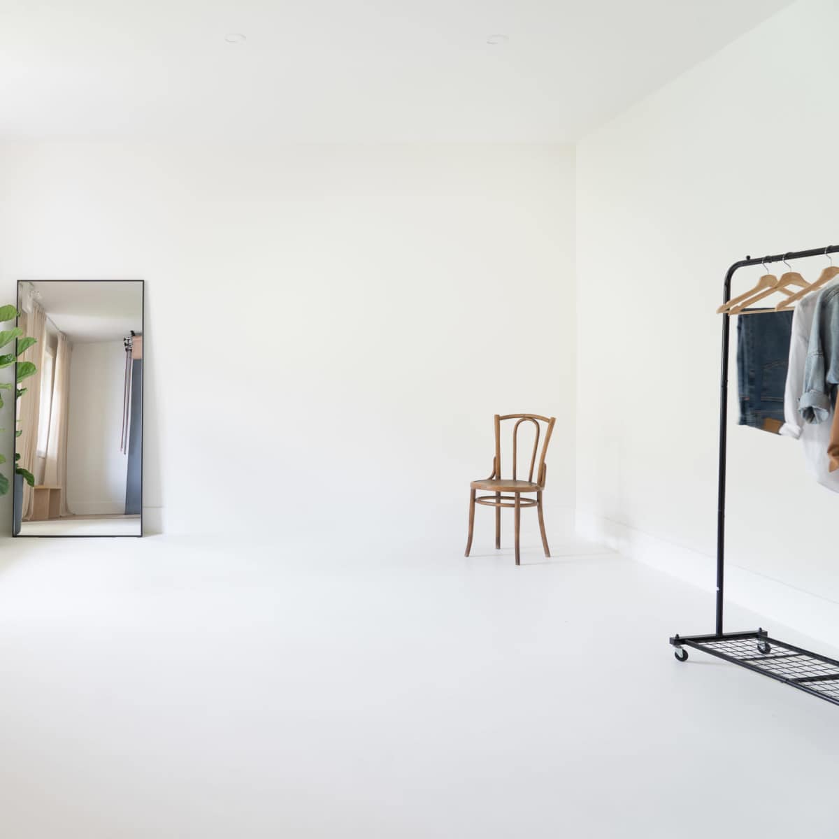 A bright, minimalist room features a tall mirror, a bench, a potted plant, a metal clothing rack with garments, and a wooden chair.